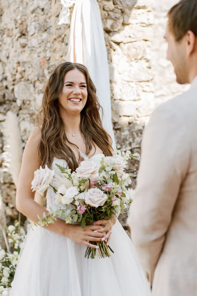A joyful bride holding a bouquet of pastel roses and greenery, smiling at her groom during their wedding ceremony.