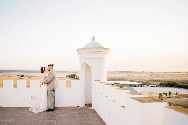 A newlywed couple embracing on a historic white castle terrace, overlooking golden fields at sunset.
