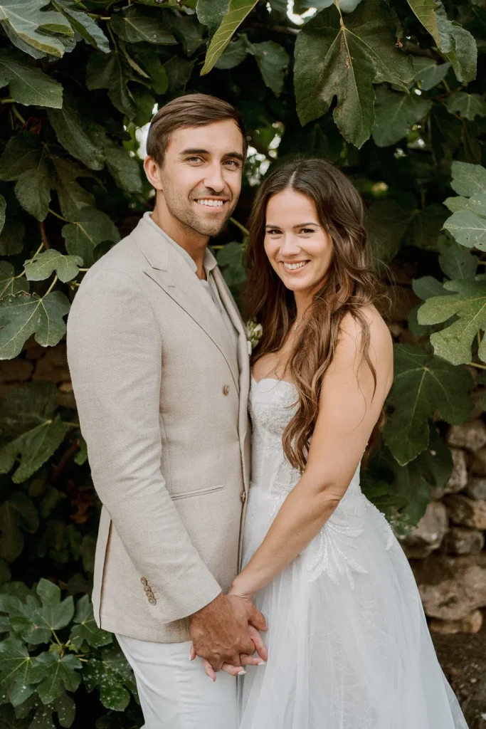 Bride and groom standing together, holding hands and smiling in front of lush green foliage.