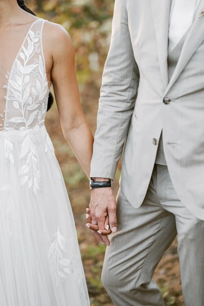 A close-up of the bride and groom holding hands, showcasing intricate lace detailing on her dress and his elegant watch.