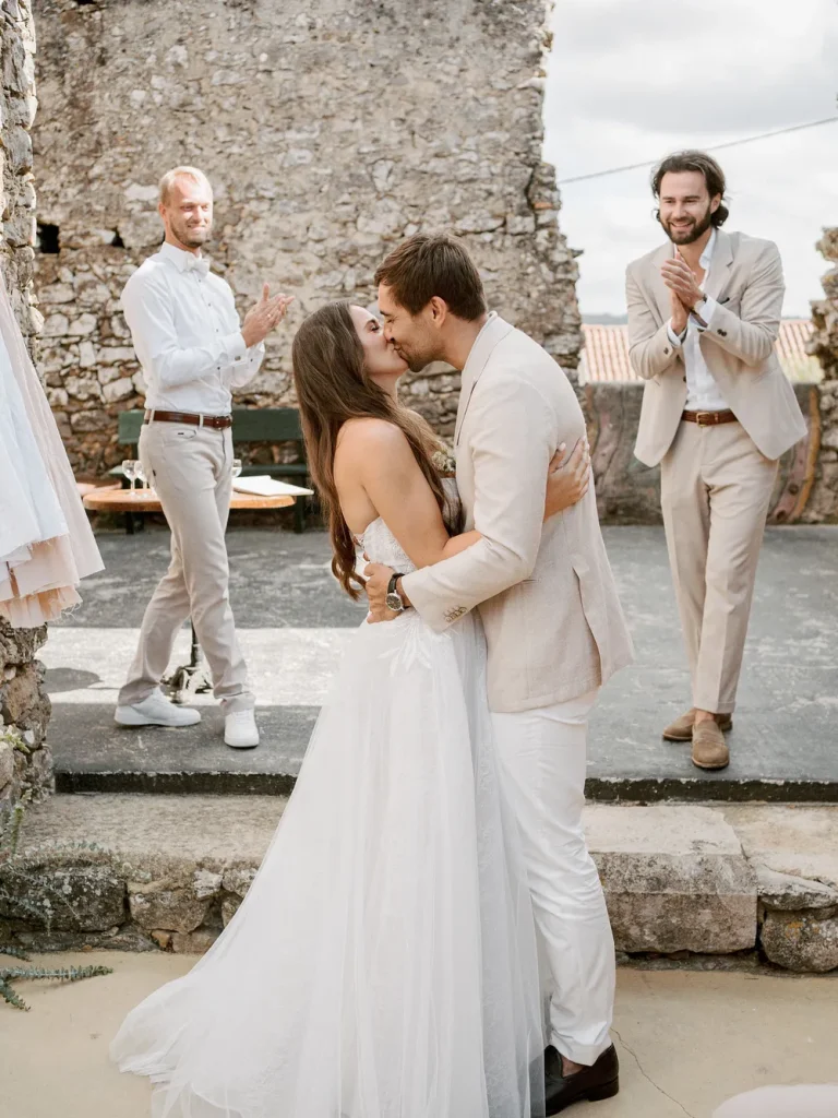 A newlywed couple shares their first kiss in a rustic outdoor wedding ceremony, surrounded by friends and family.