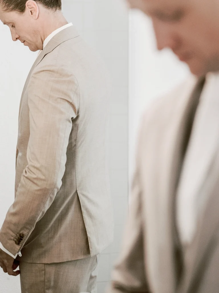 Groom adjusting his suit in front of a mirror, with a blurred figure in the foreground.