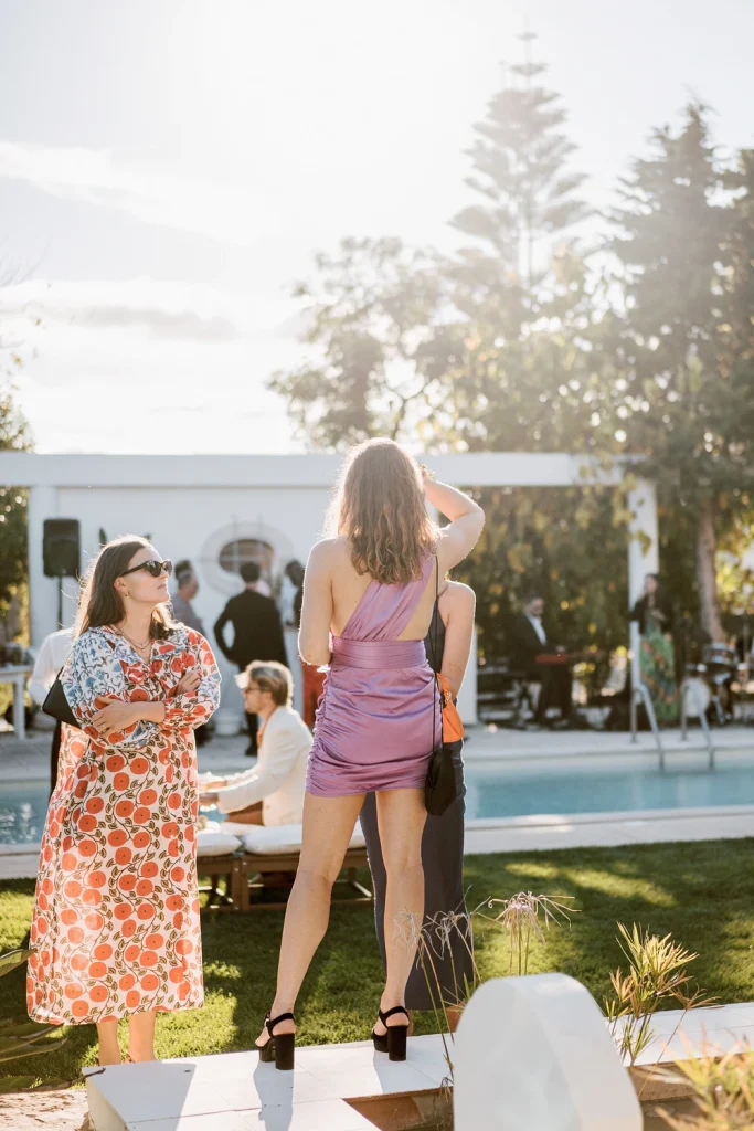 Wedding guests socializing by the poolside at sunset, dressed in stylish outfits, with live music in the background.