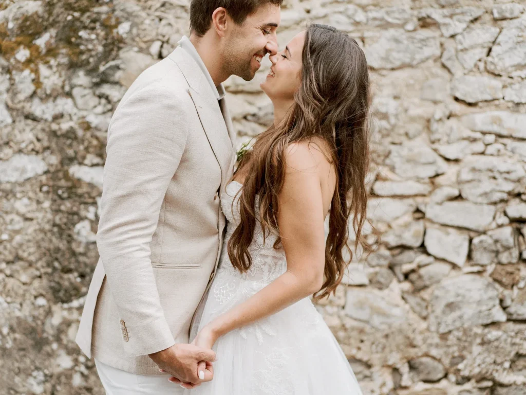A bride and groom stand close, smiling at each other with their hands intertwined against the backdrop of a textured stone wall, capturing a tender wedding moment.