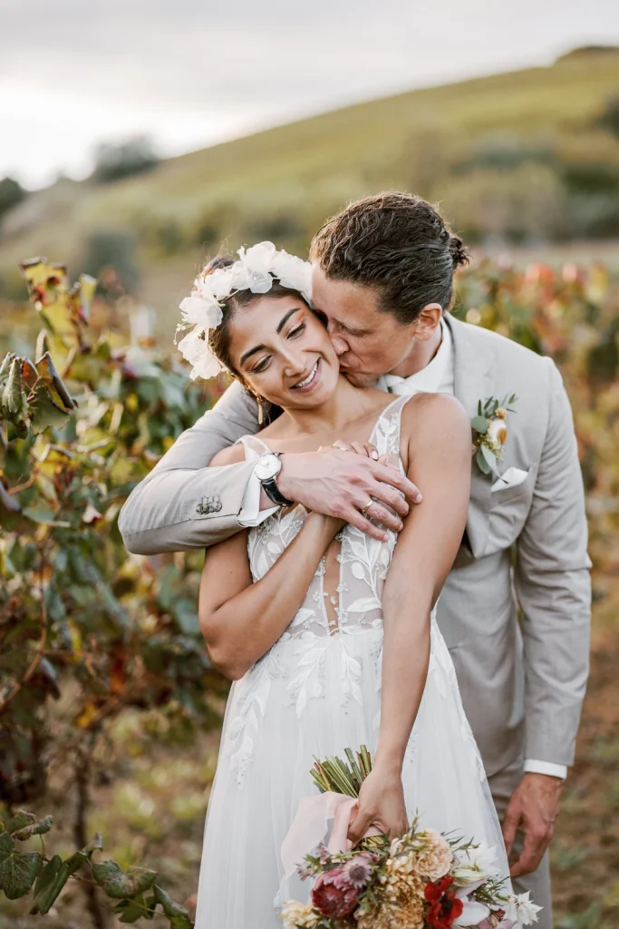 The groom lovingly embraces his bride from behind, kissing her shoulder as she smiles in a vineyard setting.