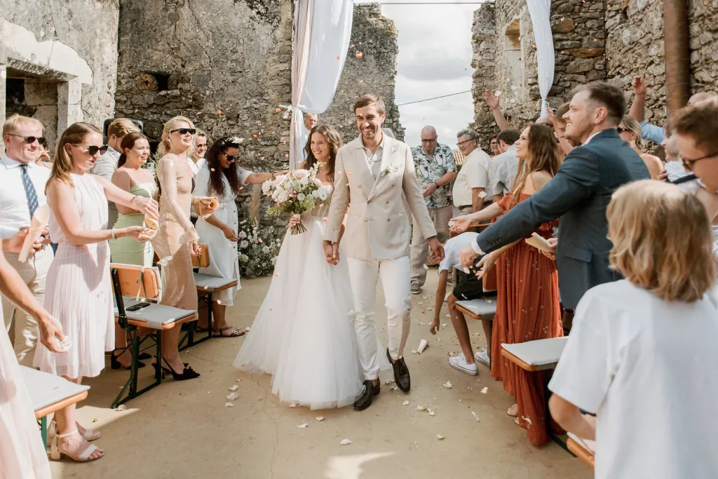 A newlywed couple walks hand in hand down the aisle as guests throw flower petals in celebration at an outdoor wedding in historic ruins.