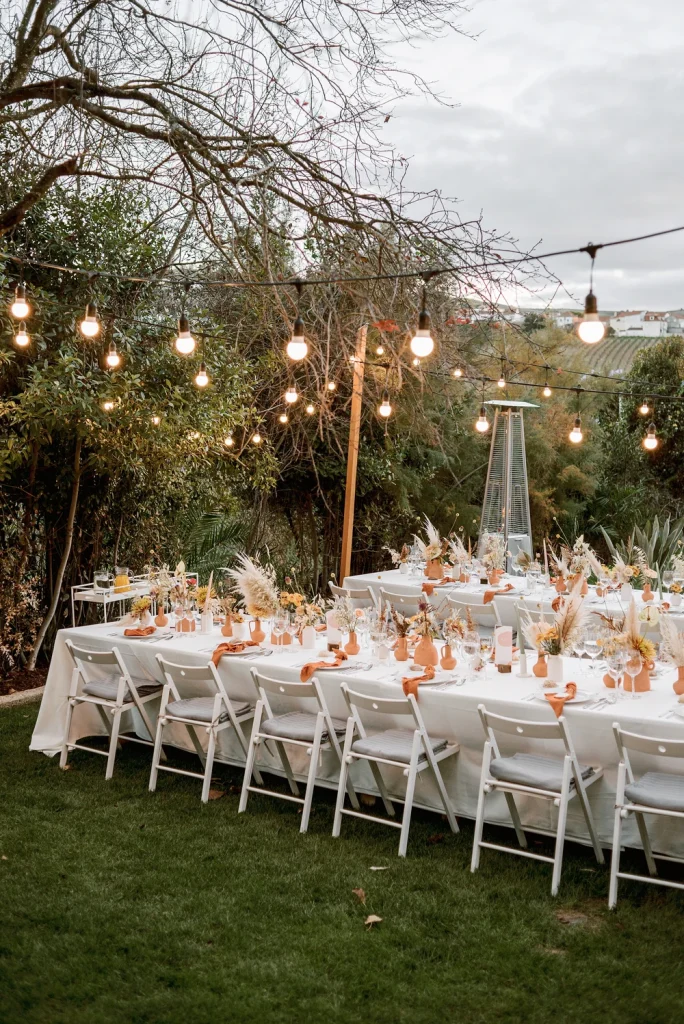 An elegant outdoor wedding reception setup with string lights, white table settings, and earthy floral arrangements.