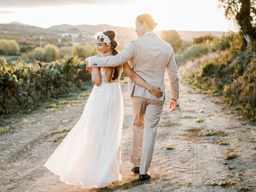 Newlyweds walking hand in hand through a vineyard at sunset, the bride's flowing dress catching the light.