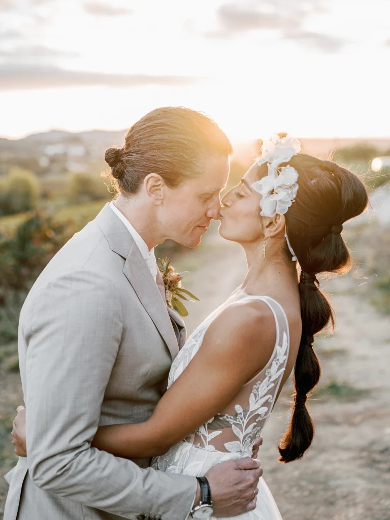 A newlywed couple shares a tender kiss in a vineyard at sunset, with golden light illuminating their embrace.